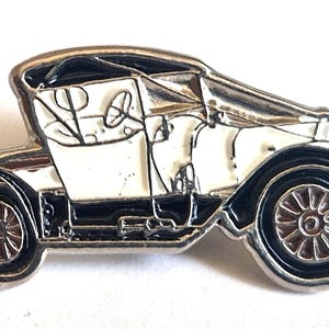 Puede incluir: Un broche de metal plateado con forma de automóvil antiguo. El diseño presenta detalles en esmalte negro en los guardabarros, las ruedas y el techo, con una carrocería en esmalte blanco. El volante y las ruedas de radios son visibles en el metal.