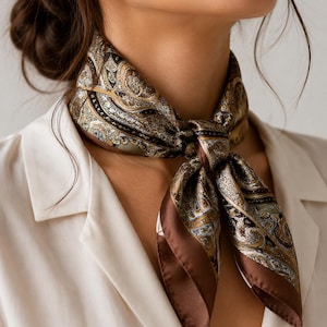 Peut inclure: Un foulard carré orné d'un motif cachemire détaillé dans des tons marron, crème et vert doux. Le foulard est noué sur le devant et présente une bordure marron foncé unie sur les bords, porté sur une chemise blanche à col simple.