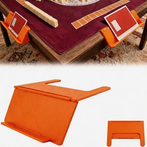 Puede incluir: Un tarjetero de plástico naranja diseñado para engancharse al borde de una mesa. El soporte cuenta con un respaldo inclinado y un borde frontal para mantener las cartas de juego en posición vertical. Se muestra en uso sobre una mesa.