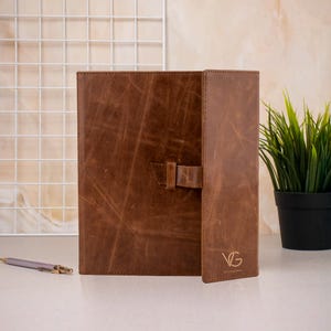 May include: A brown leather portfolio with a distressed finish and a vertical strap closure. The front flap features the embossed letters VG. The portfolio has a rectangular shape with visible stitching along the edges and a smooth, matte texture.