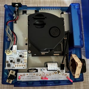 May include: An open blue translucent video game console housing with internal components exposed. Features include a black disc drive assembly, a white DreamPSU power board, a tan cooling fan, and a circuit board labelled DC Controller Board 2.0.