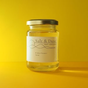 Peut inclure: Un bocal en verre transparent rempli de sirop de fleur de sureau jaune clair, doté d'un couvercle en métal argenté et d'une étiquette blanche. L'étiquette porte le texte Salt & Dune, Hand-crafted products, et Elderflower Syrup avec un dessin.