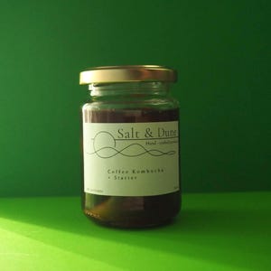 Op de afbeelding: Een doorzichtige glazen pot met een gouden deksel gevuld met een donkere vloeistof. Het witte etiket bevat de tekst Salt & Dune, Hand-crafted products en Coffee Kombucha + Starter, met een lijntekening van een zon boven een duinlandschap.