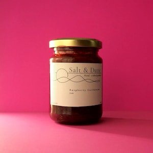 Op de afbeelding: Een glazen pot gevuld met donkerrode frambozen-kardemomjam, afgesloten met een goudkleurig metalen deksel. Het witte etiket bevat de tekst Salt & Dune, Hand-crafted products en Raspberry Cardamom Jam, samen met een lijntekening van een zon.