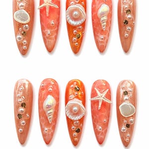 Peut inclure: Un ensemble de dix faux ongles en forme de stylet d'une teinte orange vive avec une finition texturée. Chaque ongle est décoré de petites perles blanches, de flocons dorés et de coquillages ou étoiles de mer miniatures en relief.