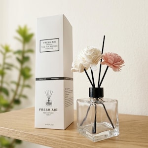May include: A clear square glass reed diffuser bottle with a black cap and three black reeds topped with two white and one pink wooden flower. Next to it stands a tall white rectangular box labelled FRESH AIR with 4 US FL OZ printed on it.