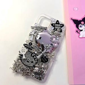 Puede incluir: Una funda de teléfono transparente decorada con varios dijes plateados, incluyendo una figura metálica de Hello Kitty, una cabeza de Hello Kitty con incrustaciones de pedrería negra, emblemas de cruces, planetas tipo Saturno y cristales.