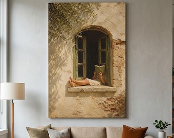 May include: A vertical wall art print depicting a person in a light-coloured garment sitting in an arched window frame with green shutters, reading an open book. The surrounding wall is textured beige stone with visible brick and olive tree shadows.
