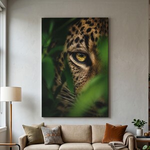 Könnte beinhalten: Ein vertikaler Wandkunstdruck mit einer Nahaufnahme eines Leopardenkopfes, der teilweise von leuchtend grünem Laub verdeckt wird. Der Leopard hat goldgelbe Augen und ein beiges Fell mit schwarzen Flecken, festgehalten in einem detailreichen Porträt.