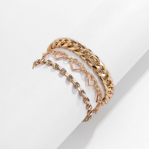 Può includere: Un set di tre bracciali color oro. Uno è una catena a maglia grossa, uno presenta una serie di maglie a forma di cuore e il terzo è una catena sottile con piccoli cristalli trasparenti a taglio rotondo disposti in una linea continua.