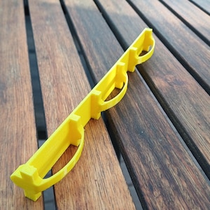 May include: A bright yellow, elongated plastic component featuring a straight central spine with three evenly spaced, curved, semi-circular loops extending from one side. The object has a rigid, structured design with a smooth, matte finish.