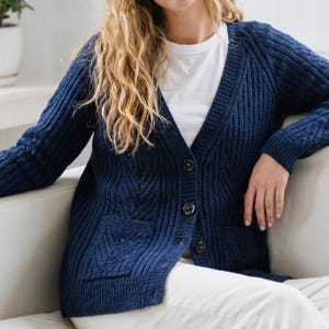 May include: A navy blue long-sleeve cardigan featuring a vertical ribbed knit pattern, a V-neckline, and three dark buttons down the front. The garment includes two front patch pockets with a chevron-style knit detail and long sleeves.