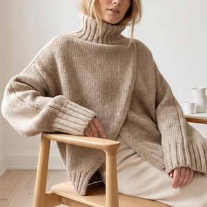 May include: A beige chunky knit sweater featuring a high ribbed turtleneck collar, long sleeves with wide ribbed cuffs, and an overlapping front panel design. The garment is crafted with a thick, textured yarn in a neutral light brown tone.
