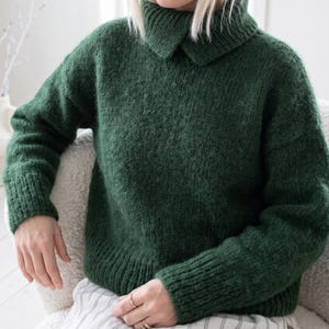 May include: A dark forest green long-sleeve jumper featuring a thick, folded-over turtleneck collar, ribbed cuffs, and a ribbed hem. The knit fabric has a textured, slightly fuzzy appearance, and the garment is constructed with dropped shoulders.