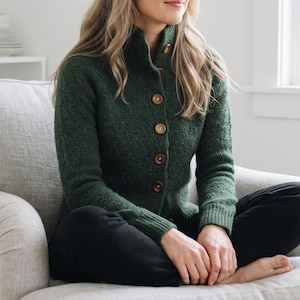 May include: A forest green knit cardigan featuring a high collar and a vertical row of five large, round, brown buttons down the front. The sweater has long sleeves with ribbed cuffs and a textured, wool-like appearance, worn over black pants.