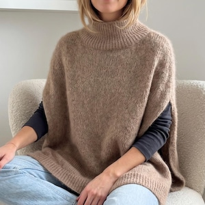 Peut inclure: Un poncho tricoté beige sans manches avec un col montant et des bords côtelés au niveau du col et de l'ourlet. Le vêtement présente une conception ouverte sur les côtés, porté sur un haut gris foncé à manches longues et un jean bleu clair.