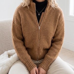 Può includere: Un cardigan in maglia color marrone chiaro, a maniche lunghe, con collo alto, polsini a coste e chiusura con zip in metallo argentato. Il capo è indossato sopra un top nero a collo alto, abbinato a una sottile collana a catena argentata.
