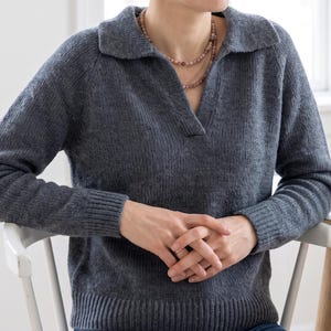 May include: A dark gray long-sleeved knit sweater featuring a collared neckline with a V-shaped opening. The garment is crafted from a textured yarn and includes ribbed cuffs and a ribbed hemline, providing a structured fit around the waist and wrists.