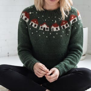 May include: A dark green long-sleeve knit jumper featuring a round neckline and a ribbed collar. The upper chest area displays a repeating pattern of small houses with red roofs and white walls, interspersed with white dots resembling falling snow.
