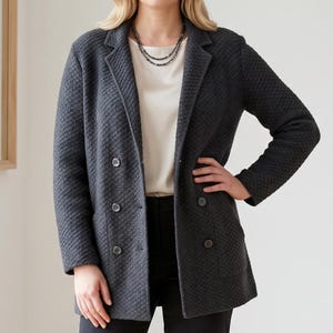May include: A charcoal gray double-breasted blazer featuring a textured, quilted-style fabric pattern, notched lapels, and four visible buttons. The blazer is worn over a plain cream-colored top and paired with black trousers, accented by a thin necklace.