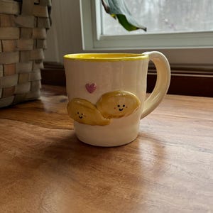 May include: Cream-colored ceramic mug with a yellow interior and handle. The mug features two smiling, yellow, cloud-like shapes and a small pink heart. The mug is sitting on a wooden surface.
