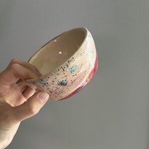 May include: A handmade ceramic bowl with a speckled pink exterior and a cream-colored interior. The bowl features a red accent on the rim and is decorated with blue and yellow accents. The bowl is held in a hand.