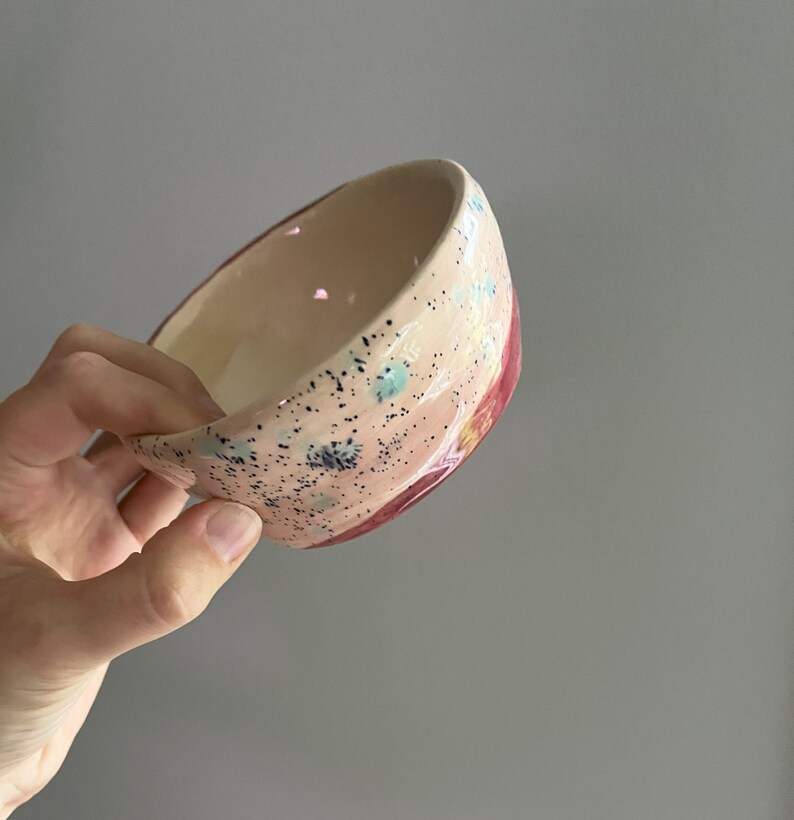 May include: A handmade ceramic bowl with a speckled pink exterior and a cream-colored interior. The bowl features a red accent on the rim and is decorated with blue and yellow accents. The bowl is held in a hand.