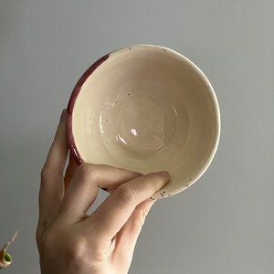 May include: A handmade ceramic bowl with a cream-colored interior and a dark red rim. The bowl has small black speckles around the rim. The bowl is held by a person's hand.