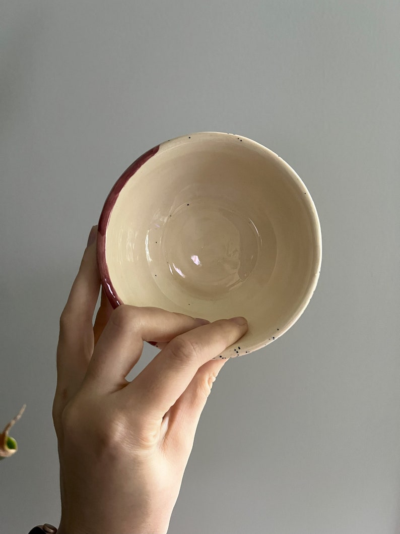 May include: A handmade ceramic bowl with a cream-colored interior and a dark red rim. The bowl has small black speckles around the rim. The bowl is held by a person's hand.