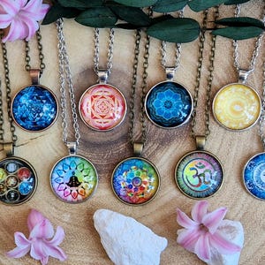 Pode incluir: Uma coleção de nove colares com pingentes redondos apresentando diversos designs espirituais e geométricos, incluindo mandalas, o símbolo Om e padrões de chakras, suspensos em correntes metálicas com engastes em tom de bronze.