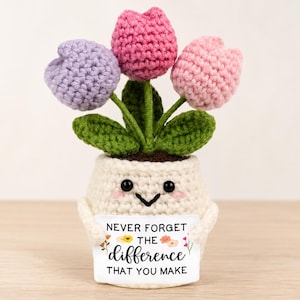 May include: A crocheted potted plant featuring three tulip-shaped flowers in lavender, magenta, and light pink with green leaves. The cream-coloured pot has a smiling face and holds a sign that reads: NEVER FORGET THE difference THAT YOU MAKE.