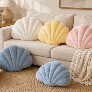 May include: A collection of five plush shell-shaped pillows in soft pastel colors including white, yellow, pink, and two blue cushions. Each pillow features a textured, fuzzy surface with stitched ridges radiating from the base to the top edge.