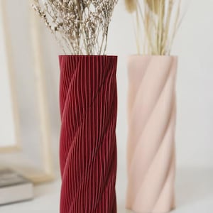 Puede incluir: Dos jarrones cilíndricos con una textura estriada y retorcida verticalmente. Un jarrón es de color rojo intenso y el otro es de color rosa pálido. Ambos jarrones están llenos de hierbas beige secas y tallos finos y delicados.