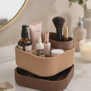 Puede incluir: Un organizador de cosméticos cuadrado de dos niveles con un exterior acanalado. El nivel superior es de color tostado claro y está dividido en varios compartimentos que contienen frascos de cuidado facial, un tubo con la etiqueta meraki y un pincel.