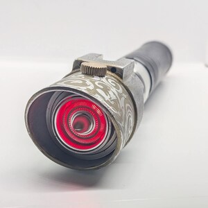 May include: A cylindrical metal flashlight with a weathered finish and a prominent red circular light emitting from the front lens. The head features an intricate etched swirl pattern and a small textured brass-colored adjustment knob on top.