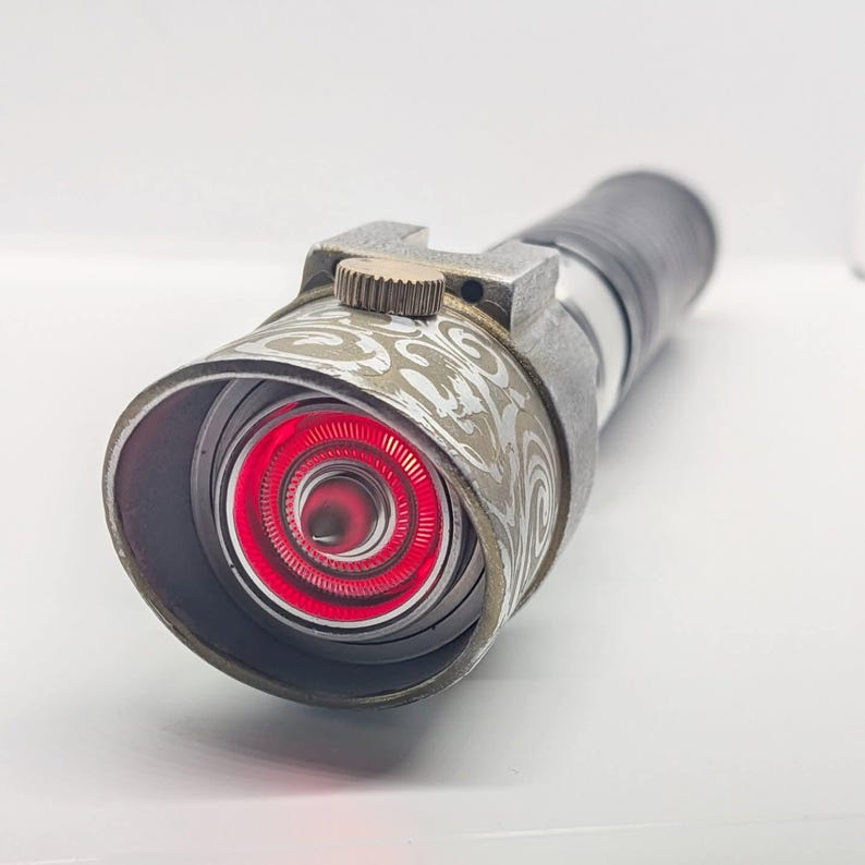 May include: A cylindrical metal flashlight with a weathered finish and a prominent red circular light emitting from the front lens. The head features an intricate etched swirl pattern and a small textured brass-colored adjustment knob on top.