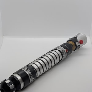May include: A cylindrical lightsaber hilt featuring a black and silver finish with intricate etched patterns. The handle includes a series of silver rings, a gold-toned ribbed section, a prominent red button, and a small red circular accent.