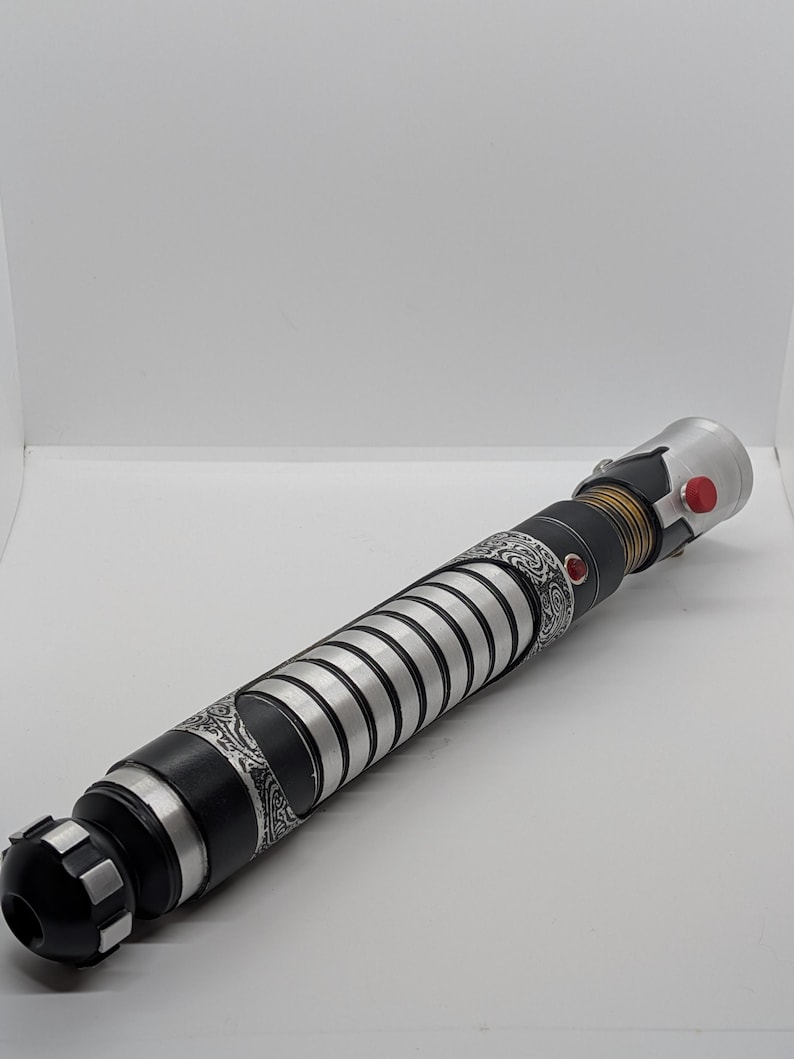 May include: A cylindrical lightsaber hilt featuring a black and silver finish with intricate etched patterns. The handle includes a series of silver rings, a gold-toned ribbed section, a prominent red button, and a small red circular accent.