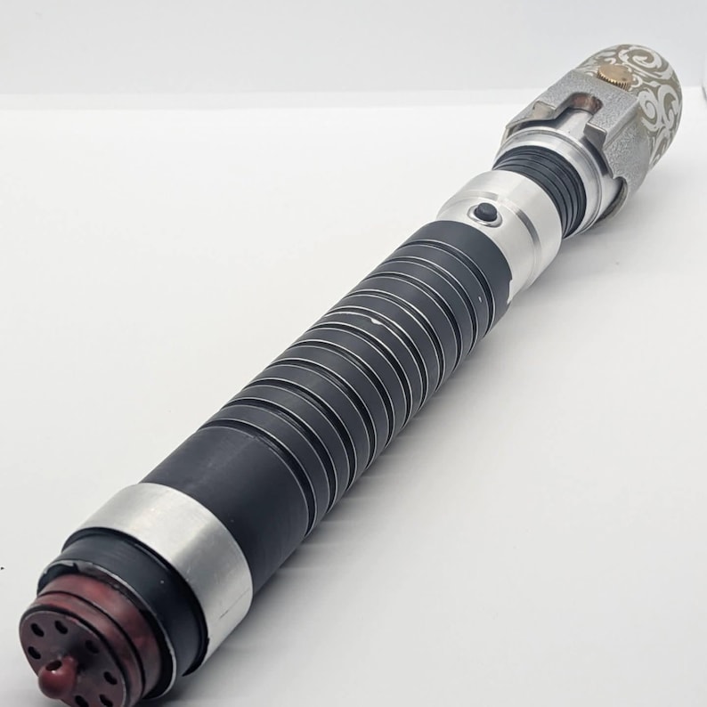 May include: A cylindrical metal lightsaber hilt featuring a black ribbed grip section, a silver emitter with a decorative white swirl pattern, a small black activation button, and a dark red circular pommel cap with multiple small ventilation holes.
