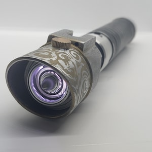 May include: A metallic prop handle featuring a cylindrical body with a textured grip and a flared emitter head. The emitter displays an intricate swirling pattern in a light tan hue, a small copper-colored dial, and a glowing purple light core.