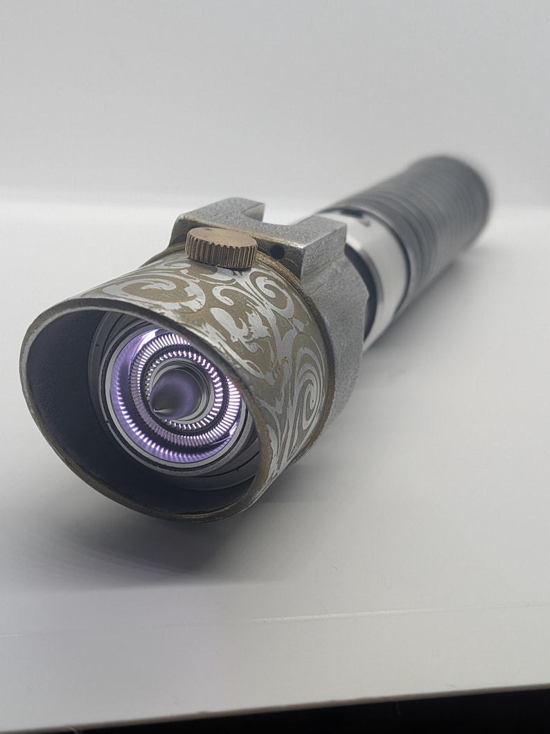 May include: A metallic prop handle featuring a cylindrical body with a textured grip and a flared emitter head. The emitter displays an intricate swirling pattern in a light tan hue, a small copper-colored dial, and a glowing purple light core.