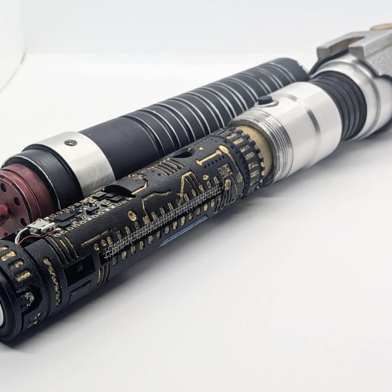 May include: A cylindrical prop weapon featuring a black handle with silver rings and a textured black grip section adorned with intricate gold-colored circuit board patterns. The device includes exposed electronic components and a metallic silver emitter.