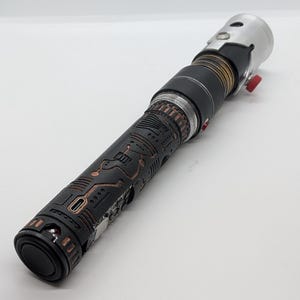 May include: A cylindrical lightsaber hilt featuring a black textured grip with copper-colored circuit board patterns. The hilt includes a silver metallic emitter, a black midsection with bronze rings, and a small red activation button on the side.