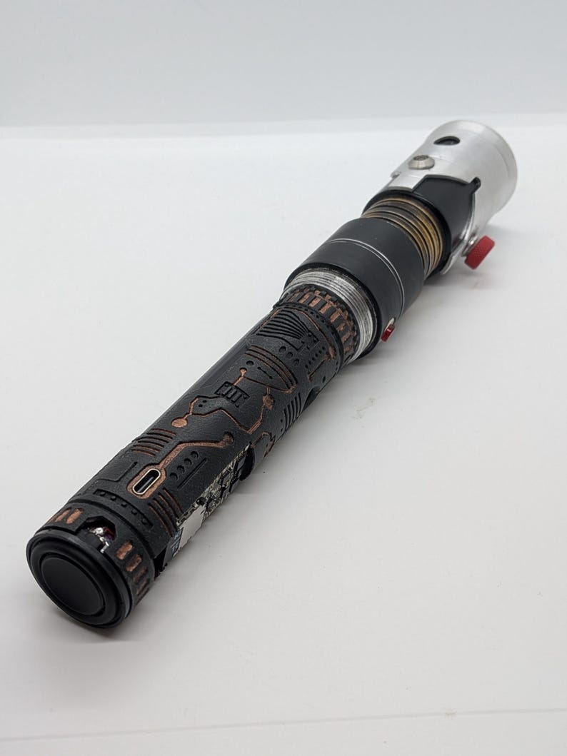 May include: A cylindrical lightsaber hilt featuring a black textured grip with copper-colored circuit board patterns. The hilt includes a silver metallic emitter, a black midsection with bronze rings, and a small red activation button on the side.