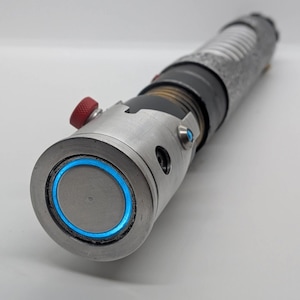 May include: A cylindrical metal prop with a weathered silver finish, featuring a prominent circular blue light ring at the emitter end, a small red textured activation button, and a black grip section with horizontal ridges along the handle.