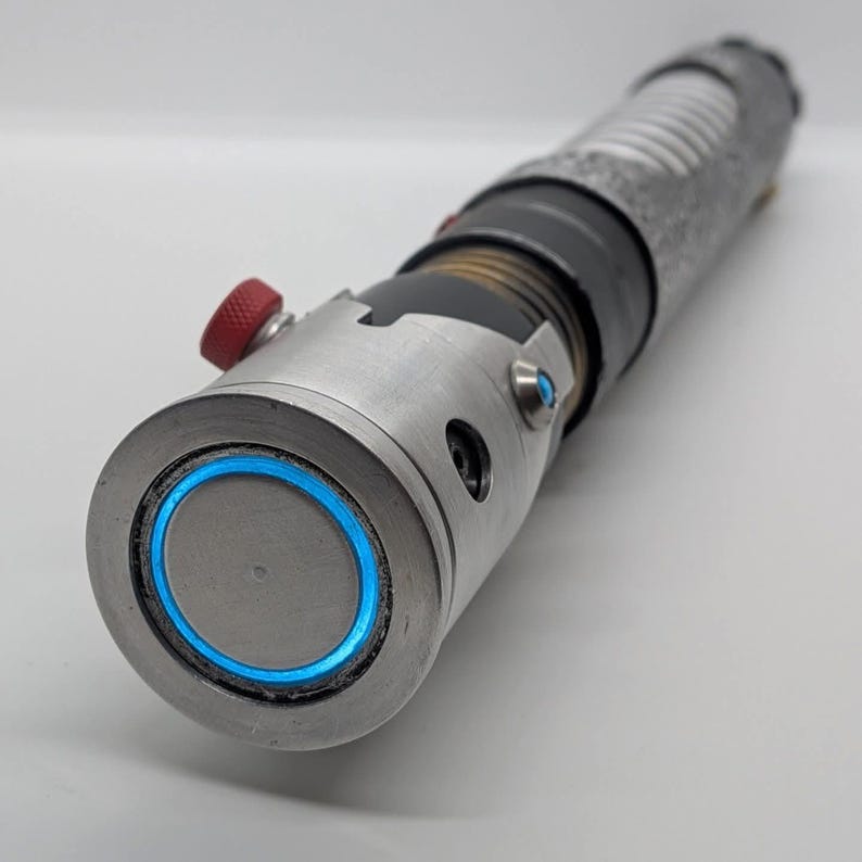 May include: A cylindrical metal prop with a weathered silver finish, featuring a prominent circular blue light ring at the emitter end, a small red textured activation button, and a black grip section with horizontal ridges along the handle.