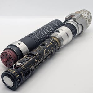 May include: A cylindrical prop with a black ribbed handle and a red circular base, positioned next to a second cylindrical prop featuring a black and gold circuit board pattern, a silver metallic section, and a white patterned top end.