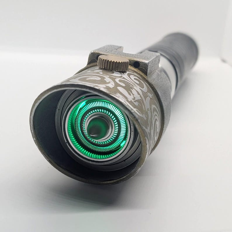 May include: A cylindrical metal prop with a textured gray finish and an intricate white swirl pattern on the emitter shroud. A small bronze-colored dial sits atop the shroud, while the interior features a glowing green circular light element.