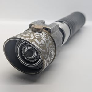 May include: A cylindrical metal prop with a weathered gray finish featuring an intricate silver swirl pattern on the emitter shroud. It includes a small brass-colored circular dial on the side and a central pointed emitter component.