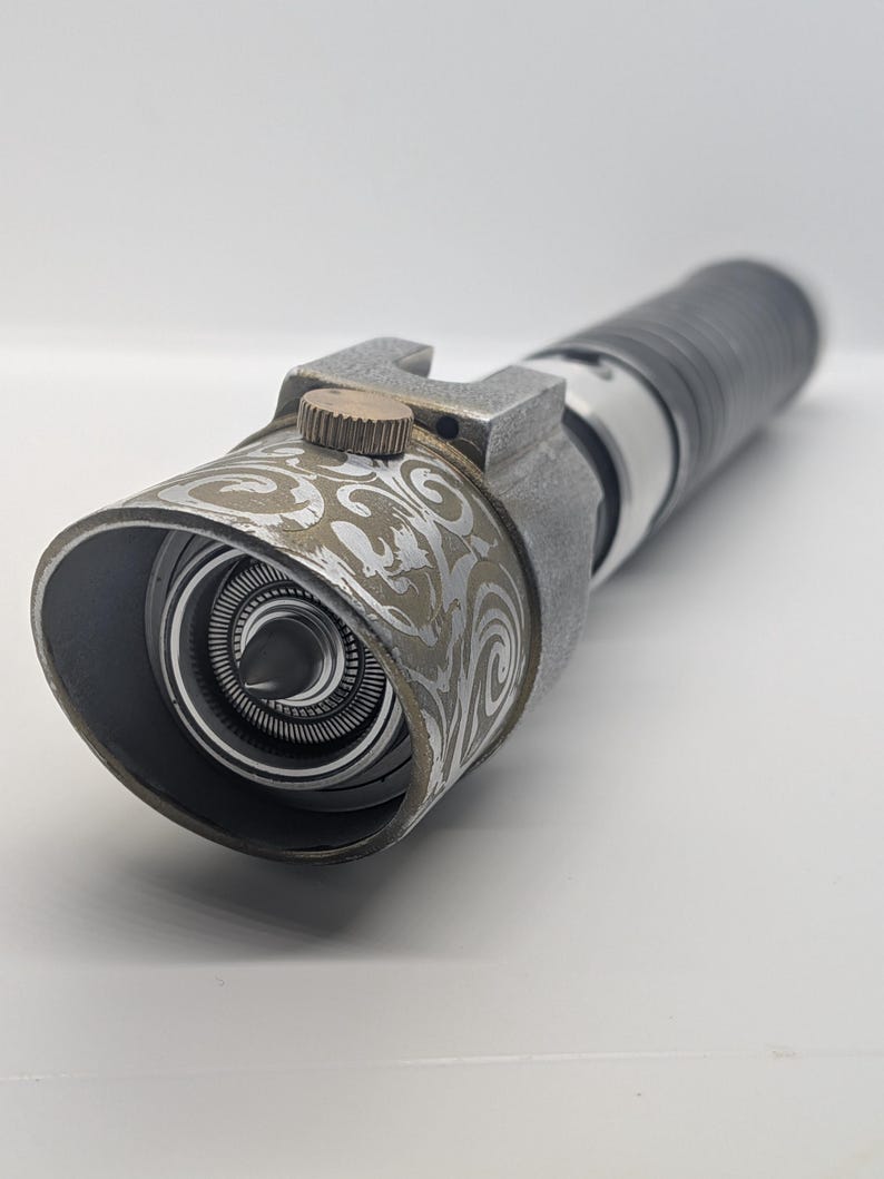 May include: A cylindrical metal prop with a weathered gray finish featuring an intricate silver swirl pattern on the emitter shroud. It includes a small brass-colored circular dial on the side and a central pointed emitter component.